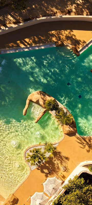 Parc aquatique vu du ciel : bassins, rochers et toboggans entourés de parasols